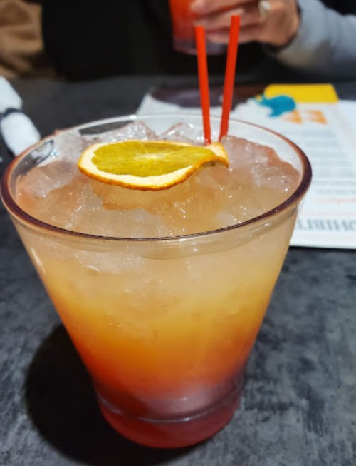 A colorful cocktail with orange slice and two red straws, served over ice in a clear glass.