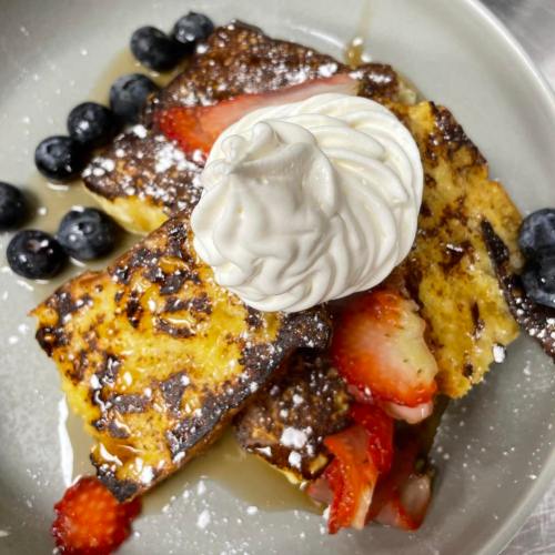 A plate of French toast topped with whipped cream, strawberries, blueberries, and a drizzle of syrup.