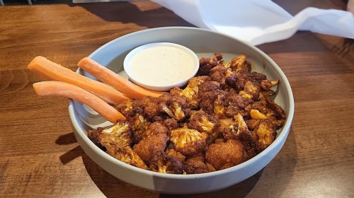 A bowl of crispy cauliflower bites with a side of ranch dressing and carrot sticks on a wooden table.