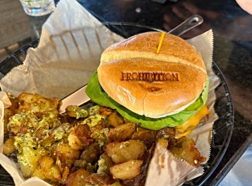 A toasted burger bun with lettuce, marked "PROHIBITION," next to a serving of crispy, seasoned potato bites.