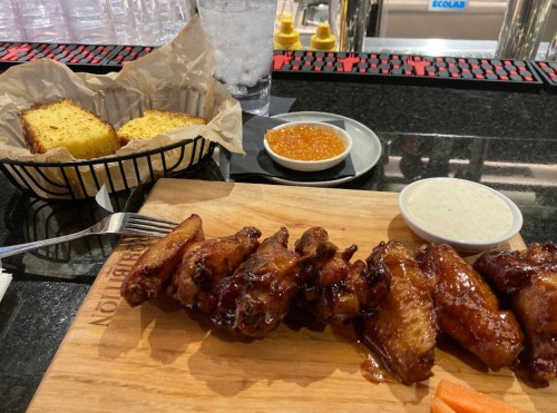 A wooden platter with glazed chicken wings, dipping sauces, cornbread, and a glass of water on a bar counter.