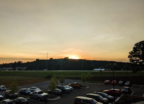 Sunset over a grassy field, with parked cars in the foreground and a silhouette of trees on the horizon.