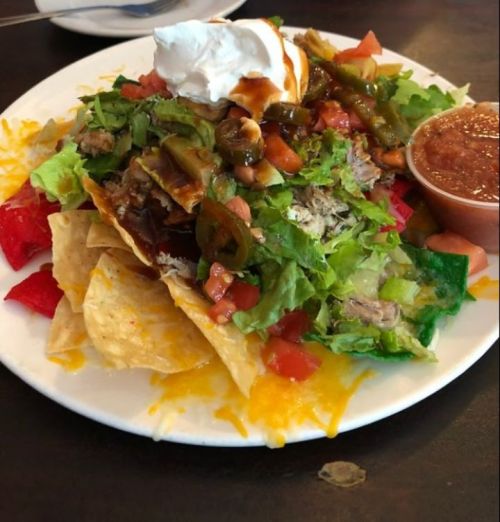 A plate of nachos topped with shredded chicken, lettuce, tomatoes, jalapeños, sour cream, and salsa on the side.