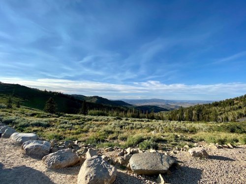 A scenic view of rolling hills and valleys under a blue sky, with rocky terrain and scattered trees in the foreground.