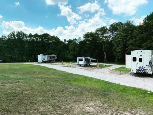 Two RVs parked on a gravel path in a green, wooded area under a blue sky with fluffy clouds.