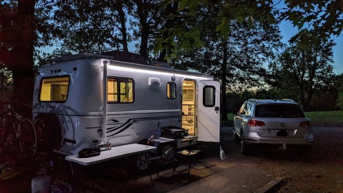 A camper trailer with lights on, parked beside a car, surrounded by trees at dusk. Tables are set up outside.