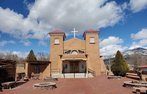 A adobe-style church with a cross, surrounded by trees and mountains under a partly cloudy sky.
