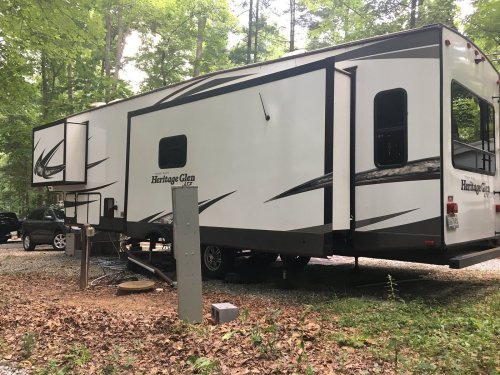 A parked RV in a wooded area, surrounded by trees and gravel, with a power hookup nearby.