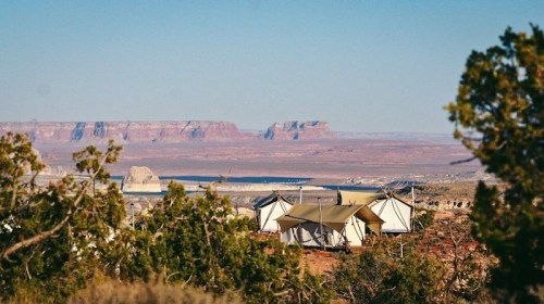 Tents nestled among trees with a scenic view of mountains and a lake in the background under a clear blue sky.