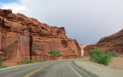 Winding road through red rock formations under a cloudy sky, with sparse greenery along the roadside.