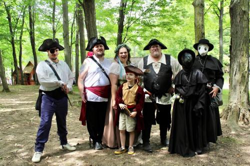 A group of seven people in costumes, including pirates and a mysterious figure, posing together in a wooded area.