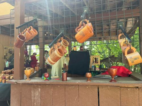 Wooden mugs and goblets hang from a display at a craft fair, with vendors and greenery in the background.