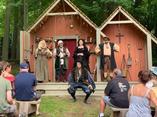 A group of performers in historical costumes entertain an audience in front of a rustic wooden stage in a forest setting.