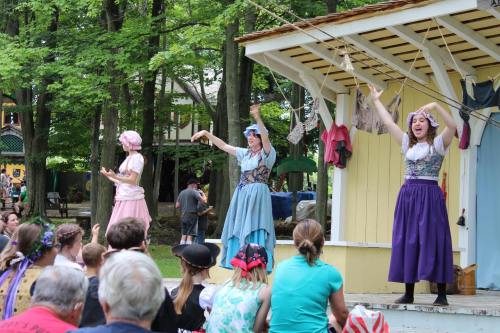 Three performers in colorful costumes entertain an audience at an outdoor event, surrounded by trees and a stage.