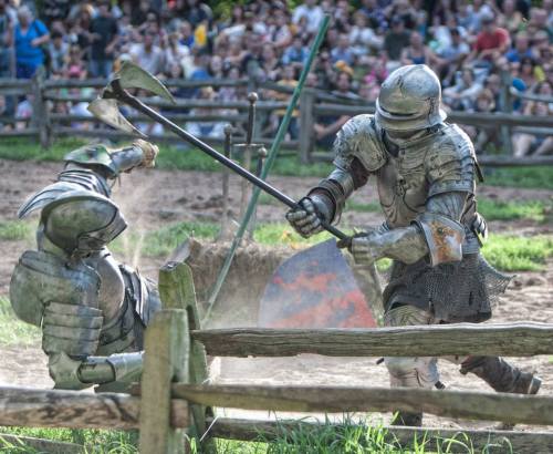 Two armored knights engage in a mock battle, wielding weapons in a dusty arena with a crowd in the background.