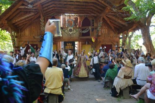 A lively outdoor festival scene with performers on stage, an audience in period costumes, and a festive atmosphere.