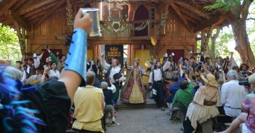 A lively medieval festival scene with performers and an audience celebrating in a rustic wooden setting.