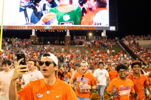 A crowd of excited fans in orange shirts celebrate at a stadium, with one person taking a selfie.