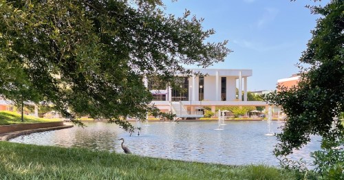A serene pond with fountains, surrounded by greenery and a heron, with a modern building in the background.