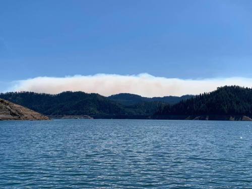 A serene lake surrounded by forested hills, with a faint haze of smoke in the distance under a clear blue sky.
