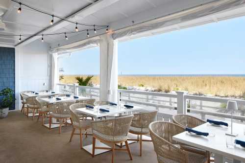 Bright coastal restaurant with white tables, woven chairs, and a view of the beach and ocean through large windows.