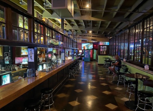 A dimly lit bar interior with a long wooden counter, shelves of drinks, and a few patrons seated at tables.