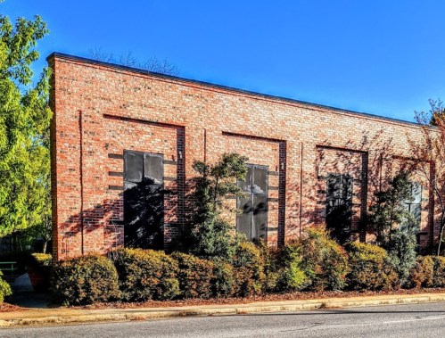 A brick building with black window shutters, surrounded by green shrubs and trees under a clear blue sky.