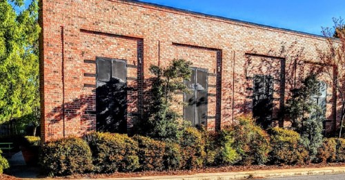 A brick building with large black windows, surrounded by green shrubs and trees under a clear blue sky.