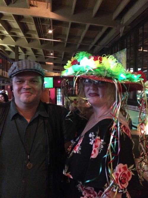 A man and woman pose together, the woman wearing a colorful, light-up hat adorned with flowers and ribbons.