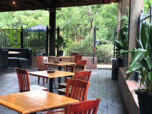Outdoor dining area with wooden tables and chairs, surrounded by greenery and a fence.