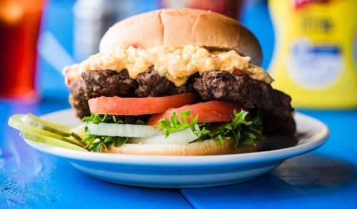 A close-up of a juicy burger with lettuce, tomato, pickles, and a creamy topping, served on a plate.