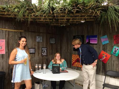 Two women and a man interact at a small outdoor gathering, with artwork displayed on the walls around them.