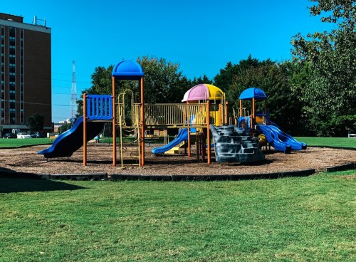 Colorful playground with slides and climbing structures, surrounded by green grass and trees under a clear blue sky.