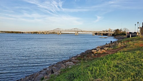 A river scene featuring a bridge in the distance, with grassy banks and a clear blue sky above.