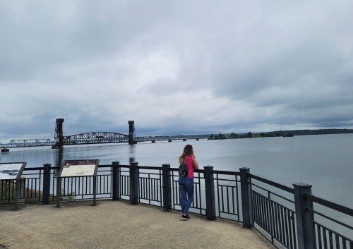 A person stands by a railing, gazing at a river and a bridge under a cloudy sky.