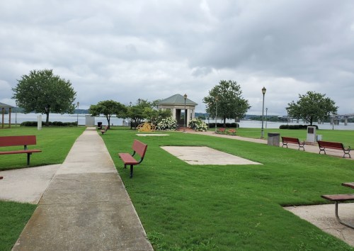 A park with green grass, benches, and a gazebo by a lake under a cloudy sky.