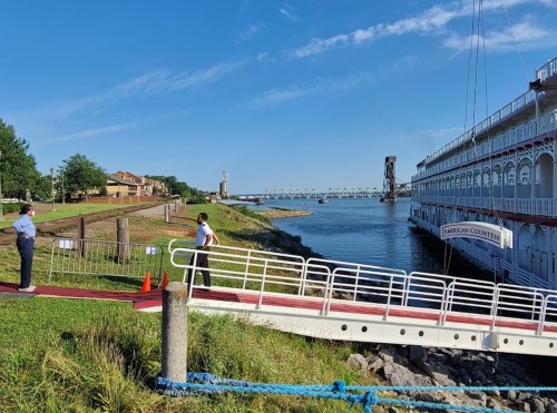 A river scene with a boat docked, two people on the shore, and a bridge in the background under a clear blue sky.