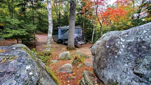 A camper parked among trees and large rocks, surrounded by autumn foliage in a serene outdoor setting.