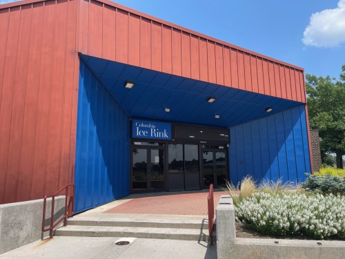 Entrance of Columbia Ice Rink, featuring a red and blue exterior with a sign above the door. Clear blue sky in the background.