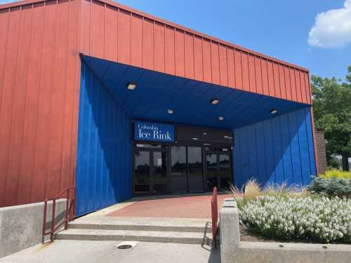 Entrance of Columbia Ice Rink, featuring a red and blue exterior with a sign above the door. Clear blue sky in the background.