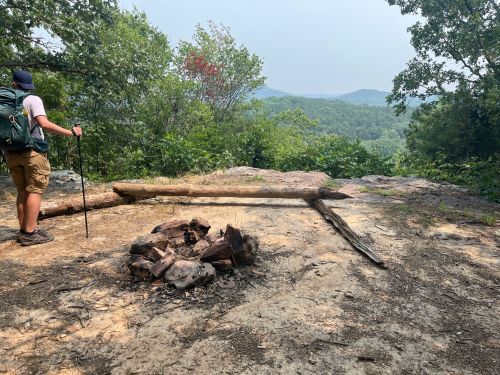 A hiker stands near a rocky overlook with a fire pit, surrounded by trees and distant mountains under a hazy sky.