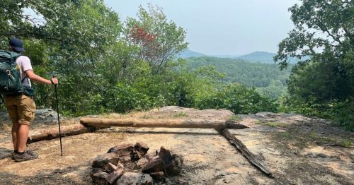 A hiker stands near a rocky overlook with a fire pit, surrounded by trees and distant mountains under a hazy sky.