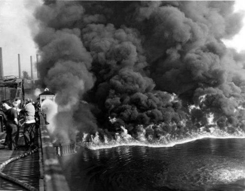 Fire and thick black smoke billow over water, with firefighters and equipment visible on a nearby dock.