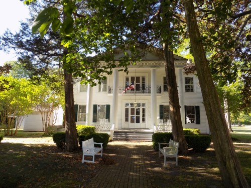 A white, two-story house with a porch, surrounded by trees and benches, set in a lush green landscape.