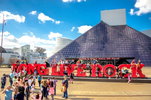 Crowd gathered outside the Rock & Roll Hall of Fame, posing with a large "LONG LIVE ROCK" sign under a blue sky.