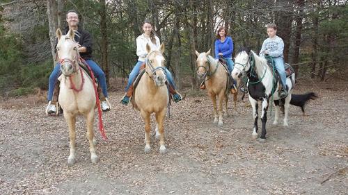A group of four people on horseback in a wooded area, with a dog visible in the background.