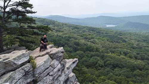 A person sits on a rocky ledge overlooking a lush green valley and distant mountains under a cloudy sky.