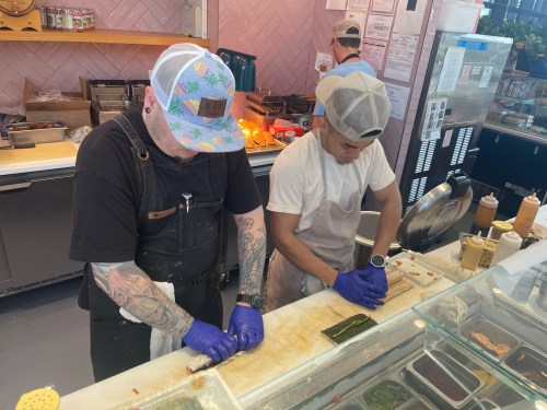 Two chefs in aprons and gloves prepare sushi rolls at a counter, focused on their work in a vibrant kitchen setting.