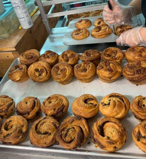 A tray of freshly baked pastries, including cinnamon rolls and pecan rolls, with hands preparing them in the background.
