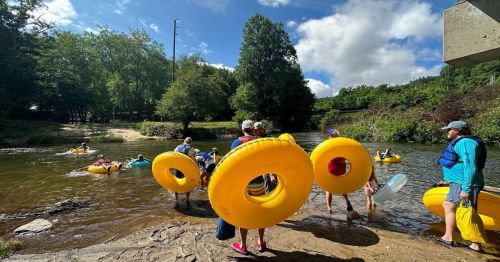 People in yellow inner tubes prepare to float down a river on a sunny day, surrounded by trees and a clear sky.